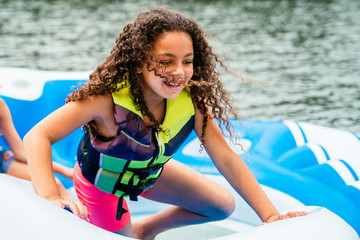 Smiling happy girl in life jacket with long curly hair playing on floaty on lake 