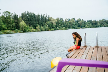 Girl in life jacket sitting on dock by lake surrounded by trees