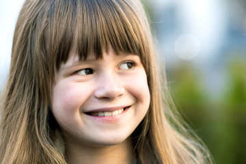 Portrait of pretty child girl with gray eyes and long fair hair smiling outdoors on blurred green bright background. Cute female kid on warm summer day outside.