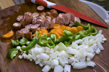 Fresh chopped vegetables and sausages  on wooden cut board with big red knife. Paprika, carrots and onion pieces. Concept of cooking process, food ingredients for saute