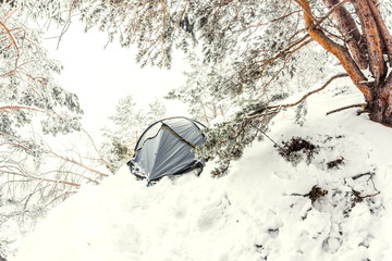 Gray tent on a dangerous uncomfortable mountainside in a snow-covered coniferous forest.