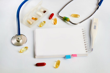 Notepad, stethoscope, electronic thermometer, colored pills and vitamins on a doctor’s table on a...