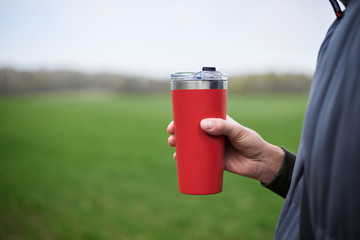 Close-up picture of male hand, holding red travel mug with hot tea coffee drink outside in the...