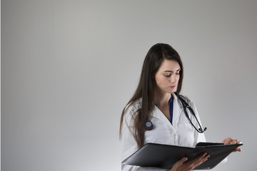 Female healthcare professional examining patient chart isolated on grey background. Stethoscope around neck, white coat on and buttoned.