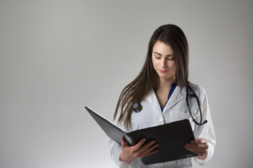 Female healthcare professional examining patient chart isolated on grey background. Stethoscope around neck, white coat on and buttoned.