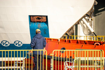Obraz premium Ferry ship ocean liner worker standing behind a yellow fence, seen from a distance, multiple huge ships in the distance. Part of a white ferry and orange tugboat