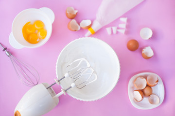 On a pink background, a hand mixer, a white bowl with cream, a whisk, eggs, eggshell, yolks on a pink background top view. Cooking, cream preparation, meringue preparation.
