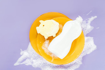 Bottle with washing agent, sponge and yellow plates on soapy foam background. Washing dishes concept. Flat lay, Top view.