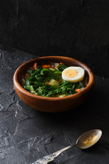 Edible wild herbs concept. Homemade rustic style soup with nettle and egg in wooden bowl with vintage fork on black background angle view