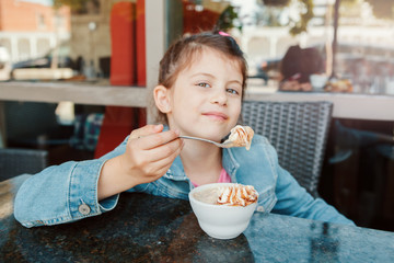 Funny Caucasian preschool girl eating sweet dessert with spoon in cafe. Child kid having fun in a restaurant patio enjoying food drink. Happy authentic childhood lifestyle.