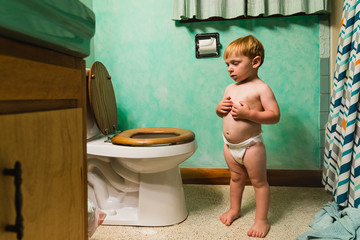 little boy in diaper stands in teal retro bathroom next to the toilet, looking at it