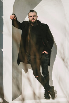 A fashionable bearded man with long hair in a black coat leans against a white wall and looks at the camera. Portrait of a fashionable guy in black clothes. Men's fashion and style.