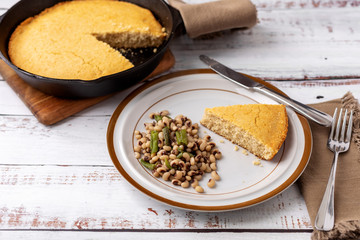 A Slice of Cornbread and Black-Eyed Peas with Snap Beans on a Brown and White Plate with a Cast Iron Skillet of Cornbread in the Background on a White Wooden Table
