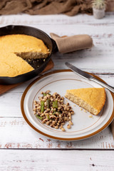 A Slice of Cornbread and Black-Eyed Peas with Snap Beans on a Brown and White Plate with a Cast Iron Skillet of Cornbread in the Background on a White Wooden Table