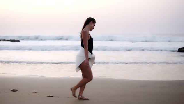 Sexy girl in a bathing black suit and white shirt tight on belly walks over a sandy beach at the ocean - gorgeous long haired caucasian girl walks alone by seaside in carefree mood, running. Side view
