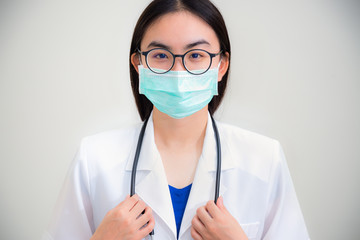 Studio portrait front face beautiful Asian young woman doctor with stethoscope in white uniform wear glasses and green mask to protect Corona Virus for health looking at camera on gray background