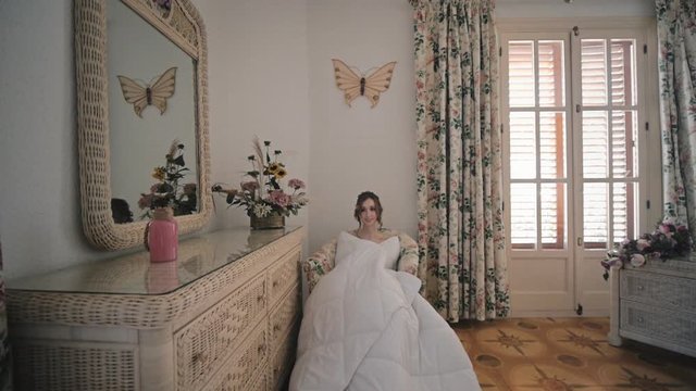 Young woman wrapped in white blanket sits in chair at home. Wide shot.