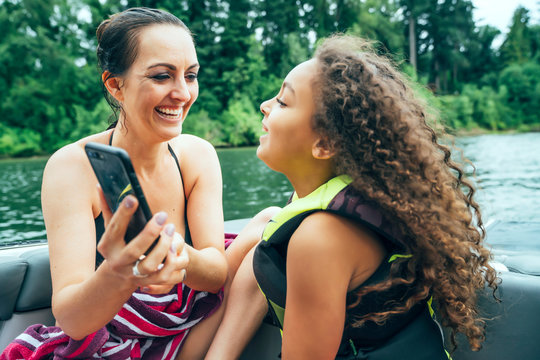 Mom and daughter laughing at video on smart phone on boat on lake surrounded by trees