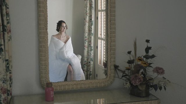 Reflection of young woman wrapped in white blanket sitting in chair. Slim girl near window at home.