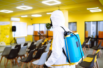 Man wearing protective suit disinfecting assembly hall with spray chemicals to preventing the...