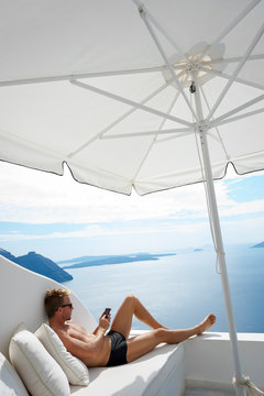 Man relaxing with his mobile phone on a white balcony with a dramatic scenic view of the Mediterranean Sea and Santorini caldera
