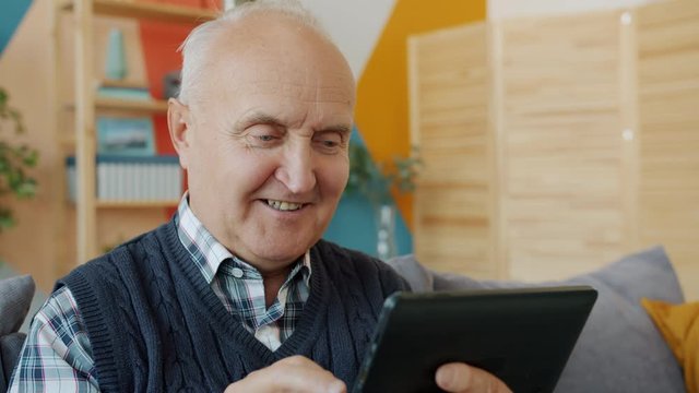 Smiling pensioner is having fun with tablet touching screen relaxing on couch at home holding modern device. Contemporary people and lifestyle concept.