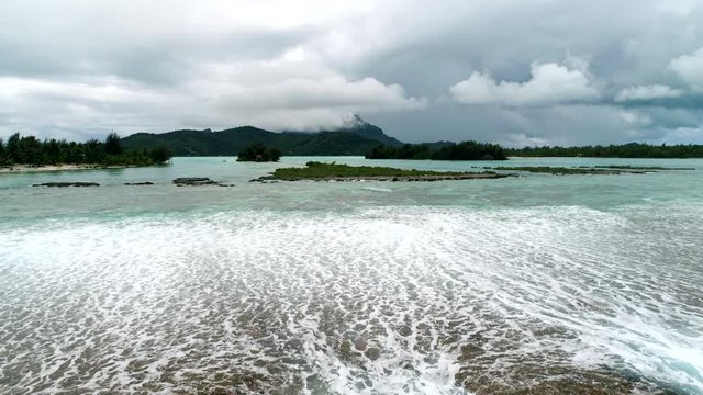 Amazing Drone shot of scenic landscape flying away from tropical lagoon reef break of paradise vacation tourist destination Bora Bora, French Polynesia. Moody rain clouds dot the background