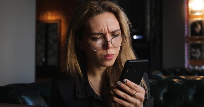 Close Up Portrait of Teen Girl with Beautiful Smile in Glasses is Using Smartphone at Day In Cafe. Casual Lifestyle. Blurred Bokeh Background. 4K