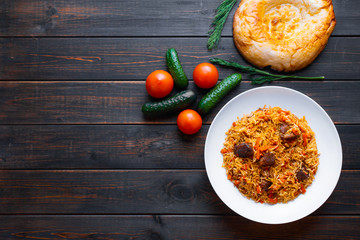 The concept of Oriental cuisine. Traditional Uzbek pilaf with lamb or beef meat on a wooden table with cucumbers, tomatoes and tandoor tortilla. View top