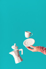 Trendy levitation Flying coffee beans, espresso cup with saucer balancing on index finger of female hand. Ceramic coffee maker levitation. Trendy mint blue coffee background, space for your text.