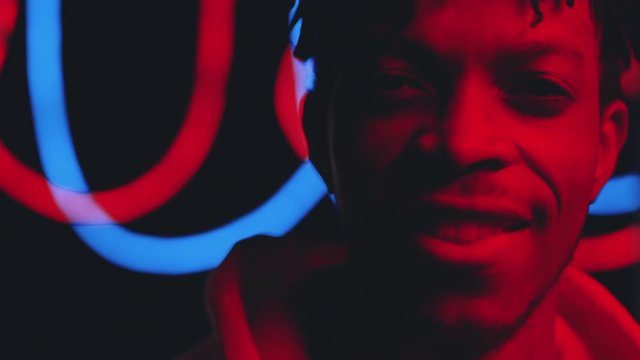 Portrait of young positive African American man looking at camera and smiling while posing in dark studio with blinking neon light and glowing multicolored tubes in background
