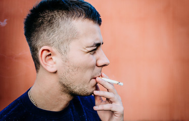 Portrait of a young man smoking a cigarette near a building wall.
