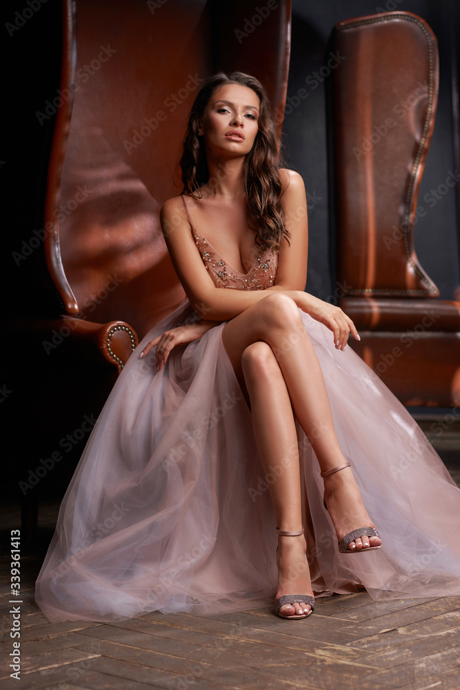 Wall mural Full length portrait of young beautiful elegant woman sitting and posing at leather chair in dark interior. Pretty female model in luxury evening dress. Lady with perfect makeup and brunette wavy hair