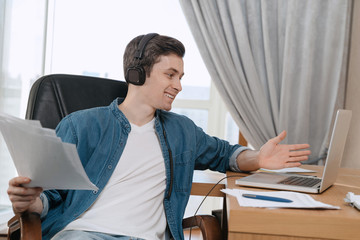Happy freelancer with headphones having conference call at home office, holding papers in his hand, gesticulating, using laptop for remote work. Young businessman has work video call, watching webinar