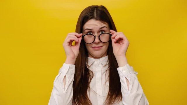 Shocked young woman keeps hands on rim of spectacles looks with omg expression gasps from shock, hears astonishing unbelievable news, wears white shirt, isolated on yellow background with copy space
