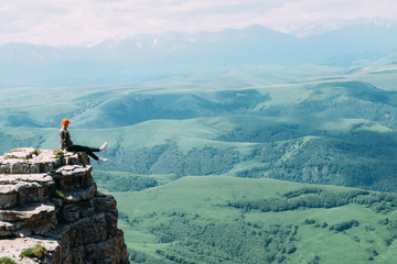 A young woman sits on the edge of a cliff against the backdrop of the Caucasus mountains and dangles her legs - 339362647