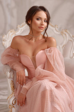 Elegant thoughtful calm woman in pink dress with decollete sitting in armchairin bright interior. Beautiful pretty caucasian woman with natural makeup and hairstyle