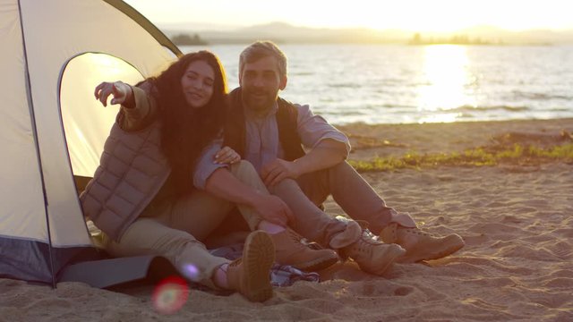 Romantic couple sitting together by camping tent on beach at sunset, smiling and pointing at something while discussing beautiful lakeshore nature