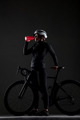 Obraz na płótnie Canvas Girl posing on roadbike. Drinking from red cycling water bottle. White protective helmet. Side lit cyclist against dark background.