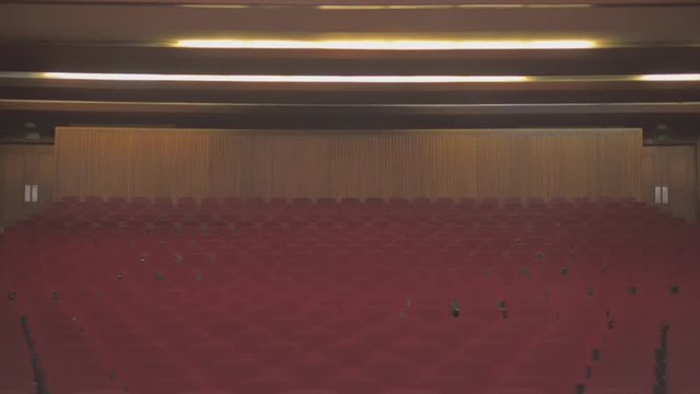 Tilt down of an abandoned theatre. The lights are on, and when the cameras gets to the end of the scene it reveals a lot of empty red chairs.