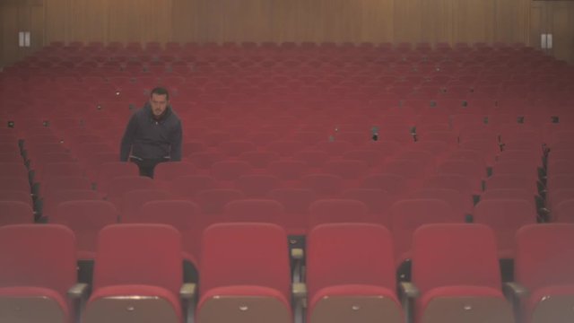 A tall white guy enters in an empty theatre. He takes a sit and he is the only one watching the show. He starts to see and enjoy the show that he sees on stage.