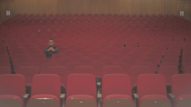 A tall white guy is in an empty theatre. He is the only one watching the show and he seems to be inspired with the performance while he pays closely attention to what he sees on stage.