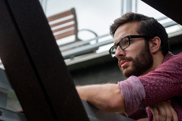 A bearded hipster man wearing a red button down shirt wearing his prescribed glasses to see more clearly what is in the distance with a straight stare on his face squinting intensely outside.