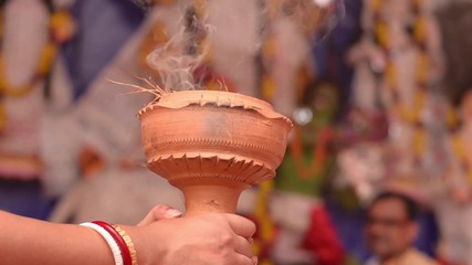 Shot of bengali woman's hands wearing red and white bangles holding Dhunachi emitting incense during pujo, used for dance and aarti in Durga Puja, with idol in the background in Kolkata, West Bengal