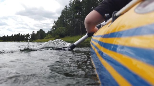 Close up view of people paddling in a raft along a river. Green pine trees are seen surrounding the river with cloudy skies.