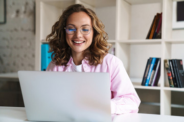 Photo of joyful woman wearing eyeglasses smiling and using laptop
