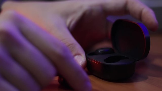 Caucasian hands in close up, place a pair of wireless earphones in their charging case which is on a wooden table. Low light, interior.