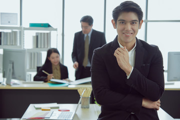 Happy asian young handsome smart successful business man smiling standing and crossed arms with team mates discussing in the background
