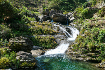 Small green waterfall, green cleat river near the green grass and forest. Rocks in the river.