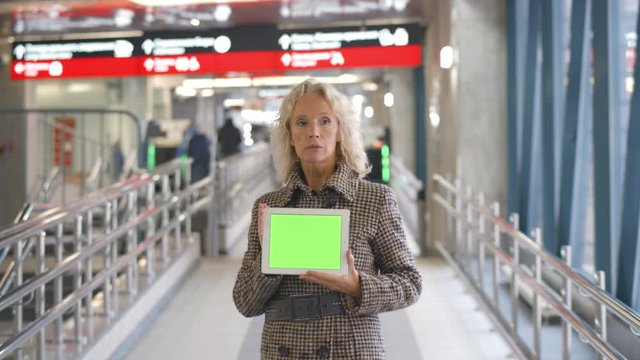 Aged Female Welcoming Guest In Airport Holding Green Screen Tablet Pc